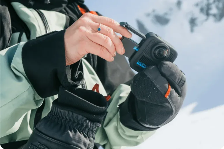 Dłoń w rękawiczce otwiera komorę baterii GoPro MAX2 na tle gór